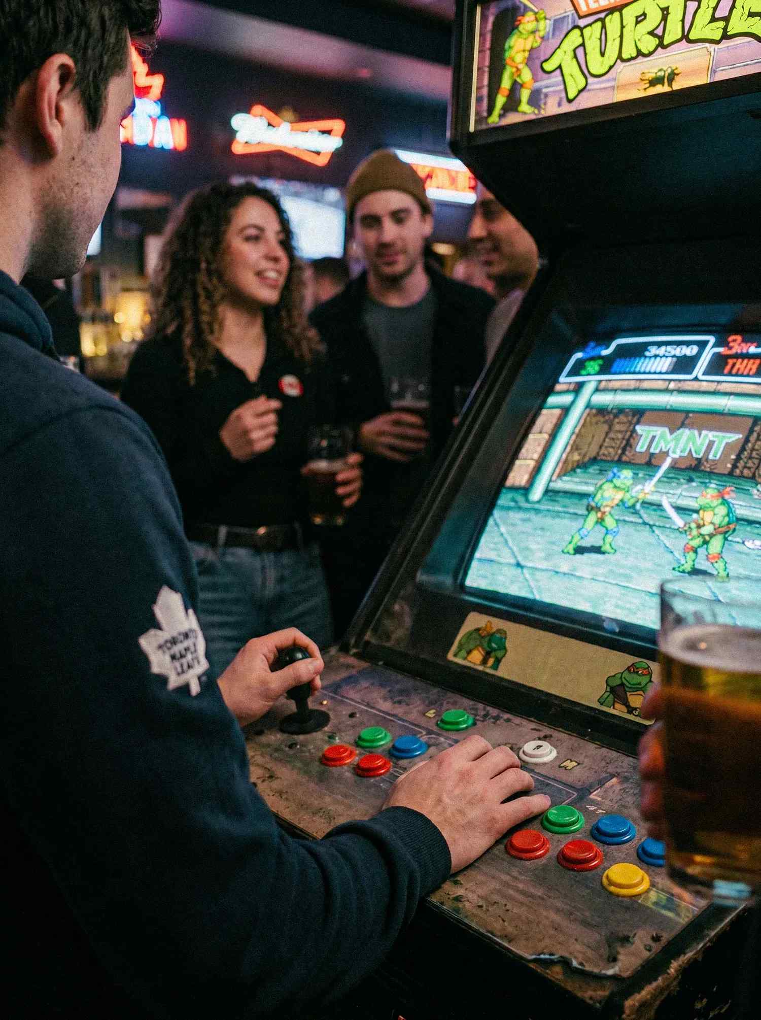 A man playing a classic TMNT arcade cabinet in a bar.